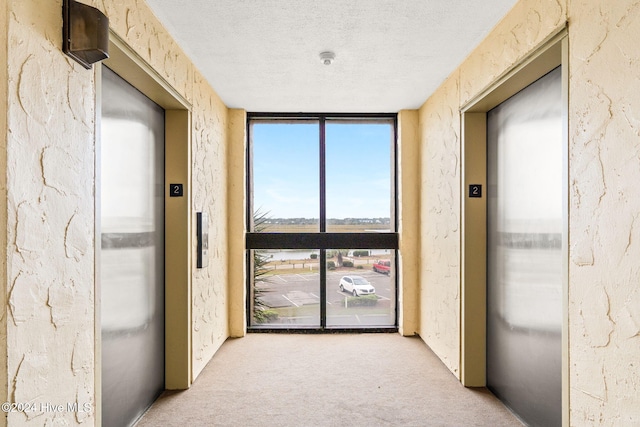 hall with light colored carpet, a textured wall, elevator, a wall of windows, and a textured ceiling