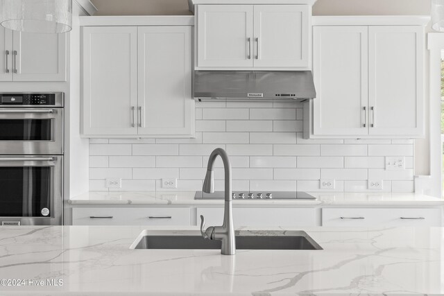 kitchen with double oven, light stone counters, white cabinetry, and exhaust hood