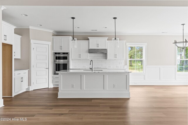 kitchen featuring white cabinets, stainless steel double oven, decorative light fixtures, and a kitchen island with sink