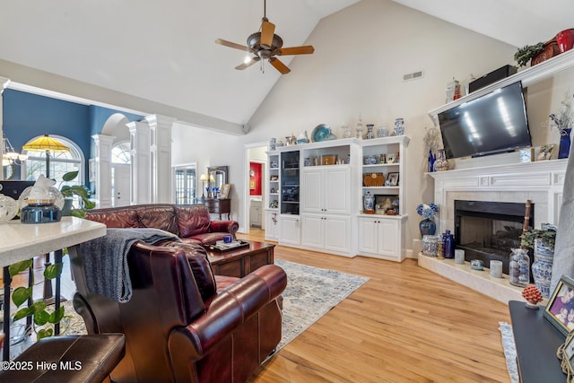 living room featuring decorative columns, ceiling fan, a tiled fireplace, light wood-type flooring, and high vaulted ceiling