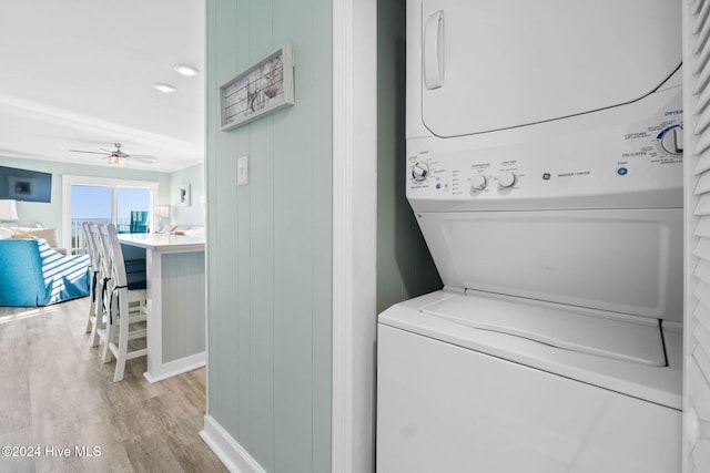 laundry area featuring ceiling fan, stacked washer and clothes dryer, wooden walls, and light wood-type flooring