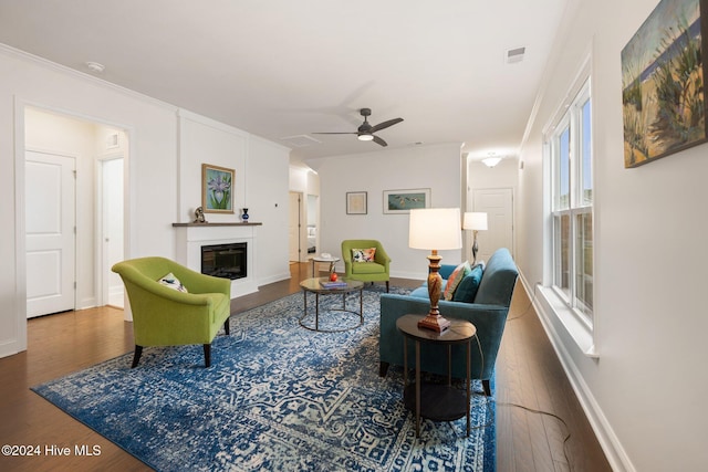 living area featuring wood finished floors, a glass covered fireplace, a ceiling fan, healthy amount of natural light, and crown molding