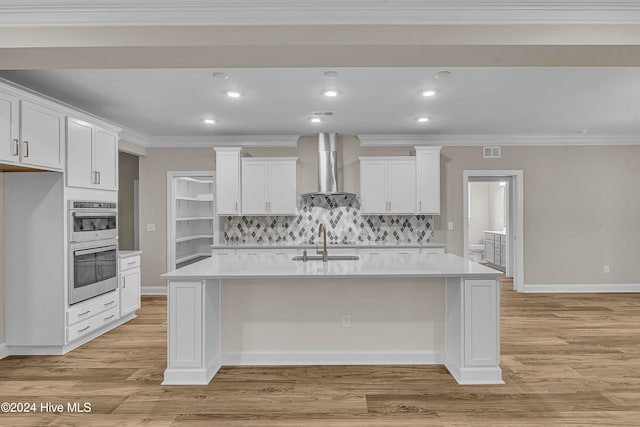 kitchen with white cabinetry, wall chimney range hood, a kitchen island with sink, and light hardwood / wood-style flooring