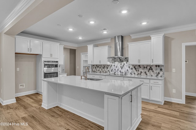 kitchen featuring double oven, sink, wall chimney range hood, a center island with sink, and white cabinetry