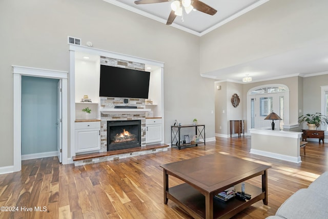 living room featuring ceiling fan, a stone fireplace, crown molding, a towering ceiling, and light wood-type flooring