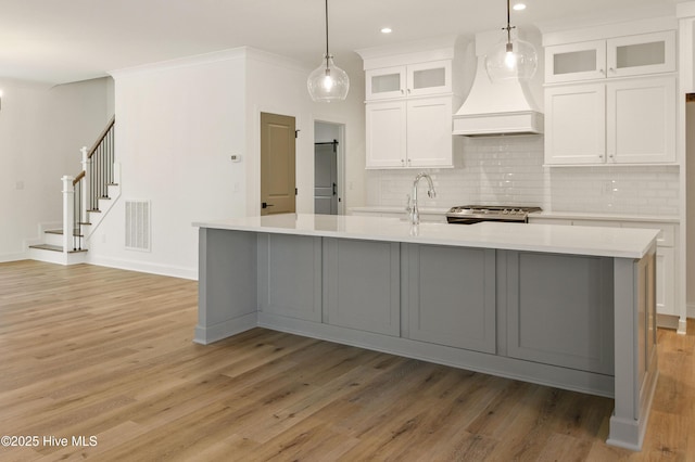 kitchen with custom exhaust hood, a center island with sink, hanging light fixtures, white cabinets, and sink