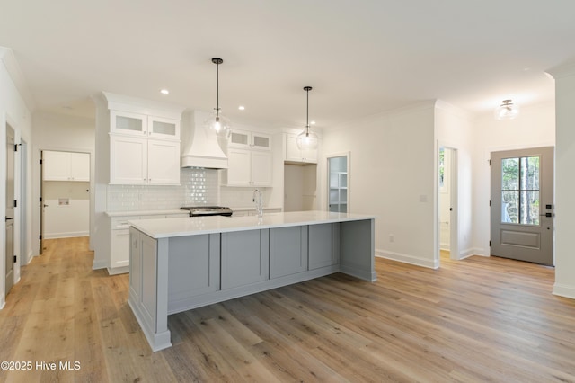 kitchen featuring white cabinets, tasteful backsplash, an island with sink, light wood-type flooring, and custom range hood