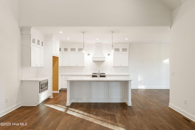 kitchen with white cabinetry, custom exhaust hood, appliances with stainless steel finishes, a kitchen island with sink, and pendant lighting