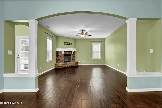 unfurnished living room featuring a stone fireplace, ceiling fan, dark wood-type flooring, and ornate columns