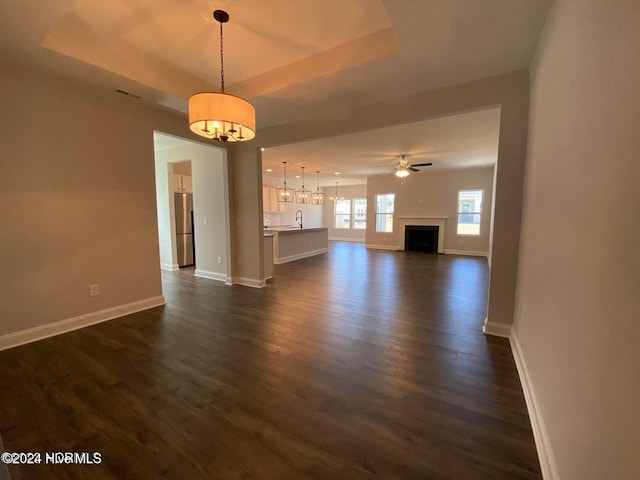 unfurnished living room with a raised ceiling, ceiling fan with notable chandelier, sink, and dark wood-type flooring