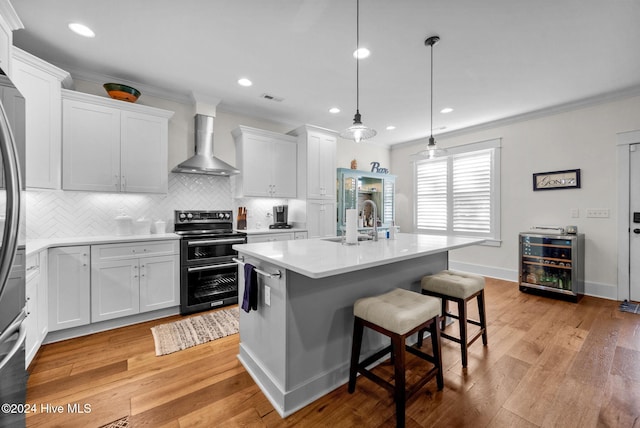 kitchen featuring electric range, wall chimney exhaust hood, an island with sink, white cabinets, and light wood-type flooring