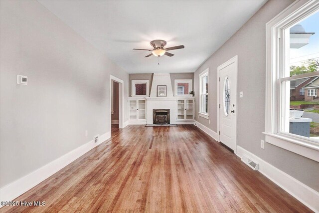 unfurnished living room featuring a ceiling fan, visible vents, a fireplace, built in shelves, and wood finished floors