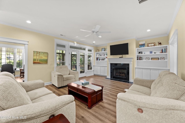 living room with a tile fireplace, ceiling fan, built in features, crown molding, and light wood-type flooring