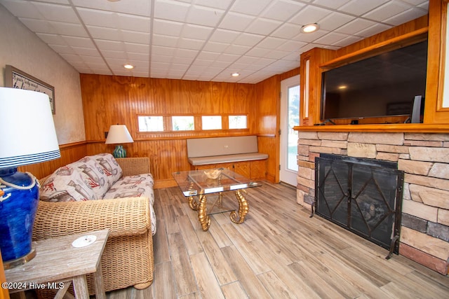 living room featuring a drop ceiling, wood walls, a stone fireplace, and light wood-type flooring