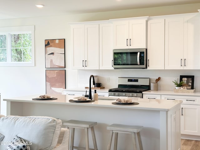 kitchen featuring appliances with stainless steel finishes, a kitchen island with sink, backsplash, white cabinets, and a kitchen bar