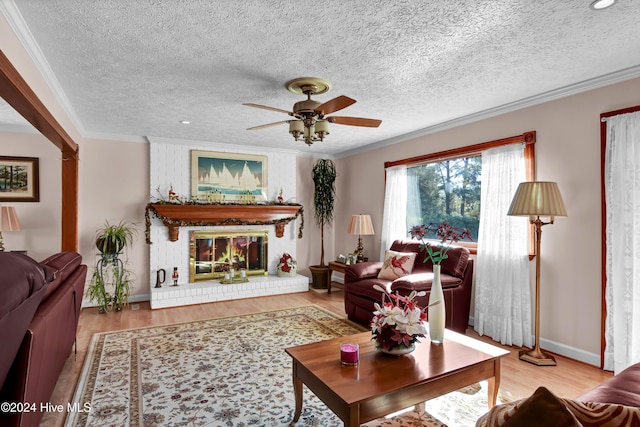 living room with ceiling fan, a brick fireplace, crown molding, a textured ceiling, and light wood-type flooring