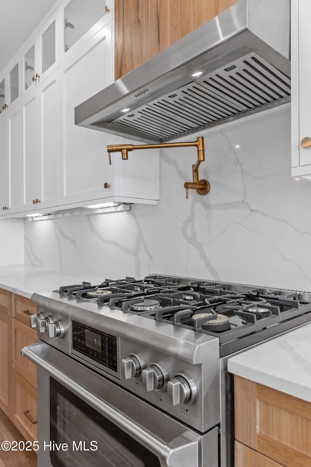 kitchen featuring stainless steel gas stove, white cabinetry, light stone counters, tasteful backsplash, and wall chimney exhaust hood