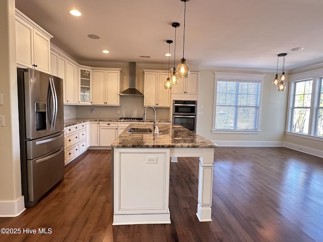 kitchen featuring an island with sink, wall chimney exhaust hood, hanging light fixtures, stainless steel appliances, and a sink