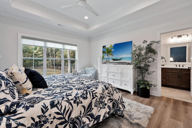 bedroom with a raised ceiling, crown molding, light hardwood / wood-style flooring, ensuite bath, and ceiling fan