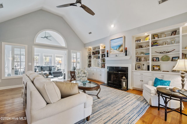 living room with high vaulted ceiling, a high end fireplace, ceiling fan, built in features, and light wood-type flooring