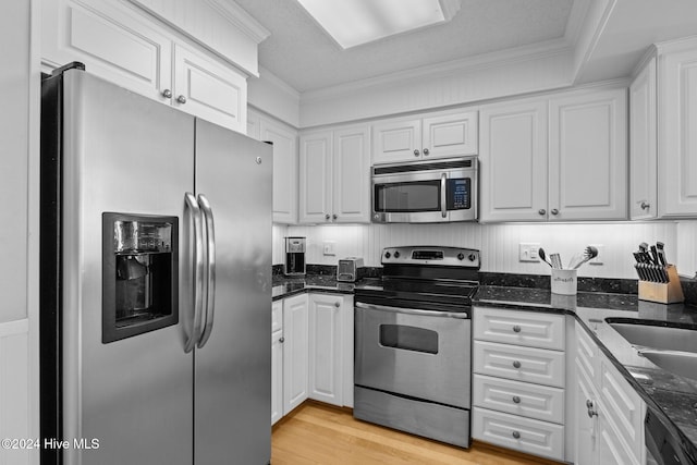 kitchen with appliances with stainless steel finishes, light wood-type flooring, dark stone counters, ornamental molding, and white cabinetry