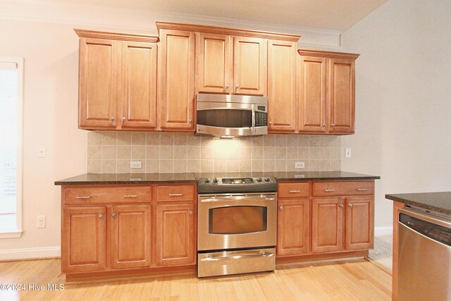 kitchen with decorative backsplash, stainless steel appliances, crown molding, dark stone countertops, and light hardwood / wood-style floors