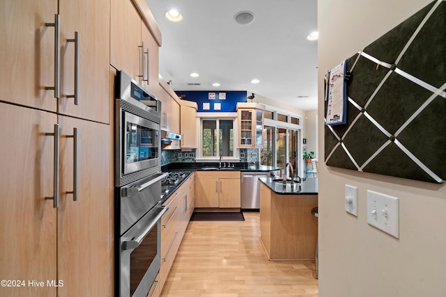 kitchen with light brown cabinetry, dark countertops, a sink, light wood-type flooring, and appliances with stainless steel finishes