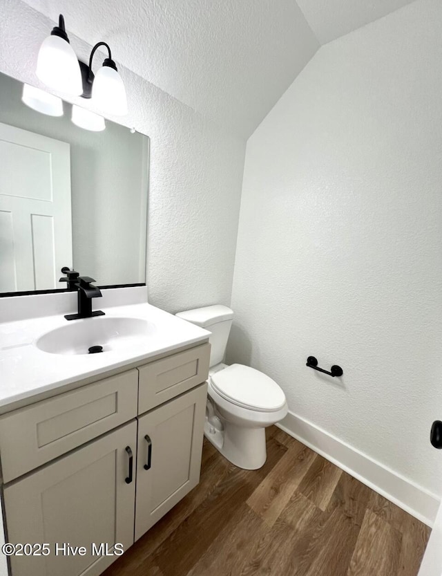 half bathroom featuring a textured wall, toilet, lofted ceiling, wood finished floors, and vanity