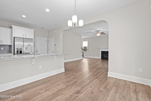 kitchen featuring stainless steel refrigerator with ice dispenser, light stone counters, ceiling fan with notable chandelier, light hardwood / wood-style floors, and white cabinetry