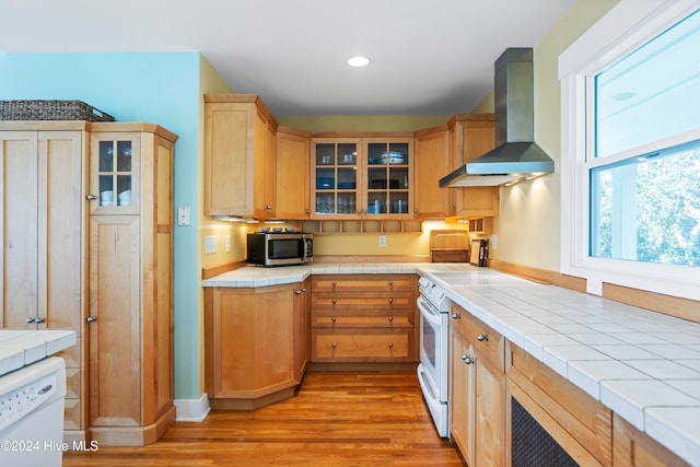 kitchen featuring tile counters, wall chimney exhaust hood, white appliances, and light hardwood / wood-style flooring