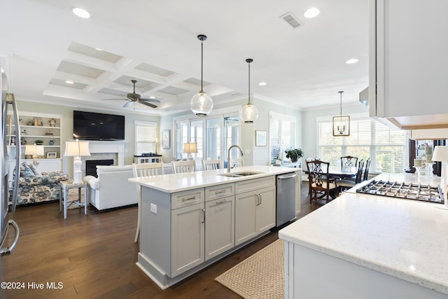 kitchen featuring coffered ceiling, stainless steel appliances, sink, hanging light fixtures, and an island with sink