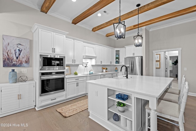 kitchen featuring appliances with stainless steel finishes, a kitchen island with sink, decorative light fixtures, beamed ceiling, and white cabinetry