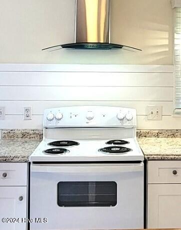 kitchen with electric range, light stone counters, white cabinetry, and wall chimney range hood