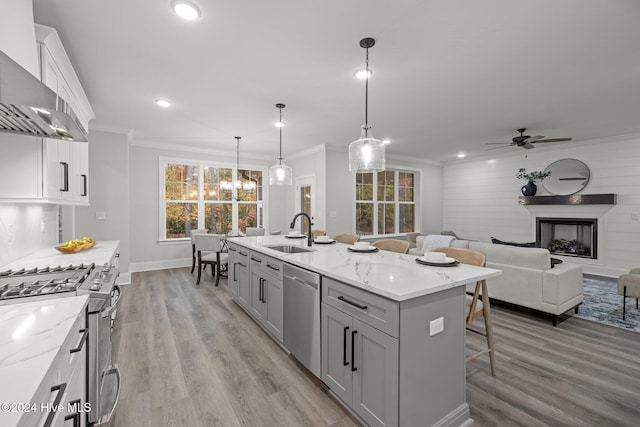 kitchen with appliances with stainless steel finishes, wall chimney exhaust hood, a kitchen island with sink, sink, and pendant lighting