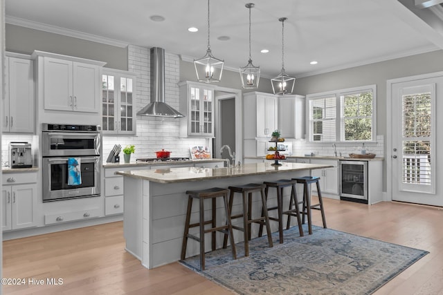 kitchen featuring a kitchen island with sink, white cabinets, wall chimney range hood, stainless steel appliances, and beverage cooler