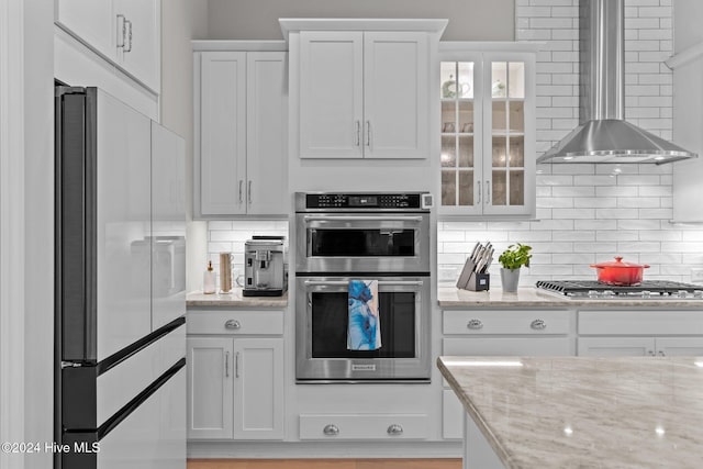 kitchen featuring wall chimney exhaust hood, light stone countertops, white cabinets, and stainless steel appliances