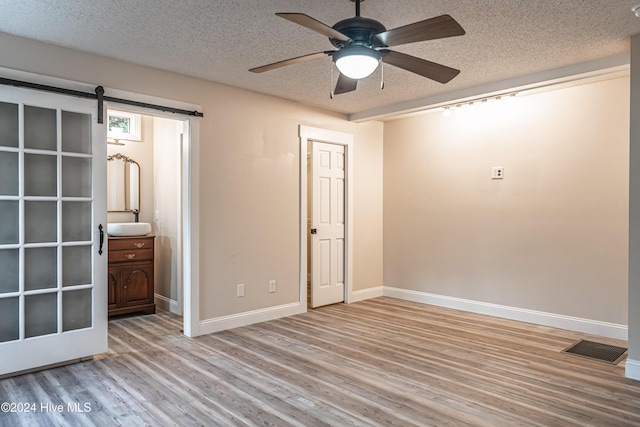 unfurnished bedroom with ceiling fan, a barn door, ensuite bathroom, light hardwood / wood-style floors, and a textured ceiling