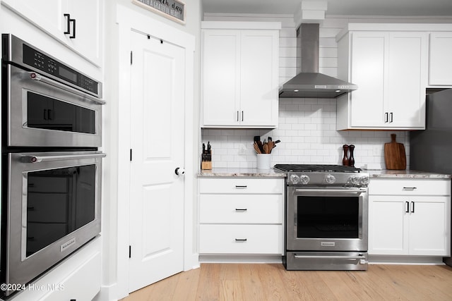 kitchen featuring wall chimney exhaust hood, light stone countertops, light wood-type flooring, appliances with stainless steel finishes, and white cabinetry