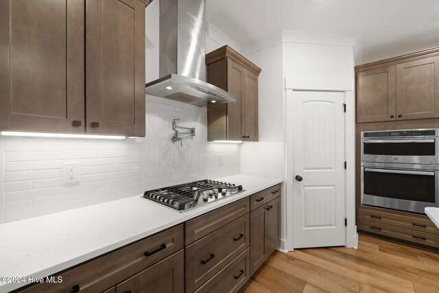 kitchen with wall chimney range hood, appliances with stainless steel finishes, light wood-type flooring, and tasteful backsplash