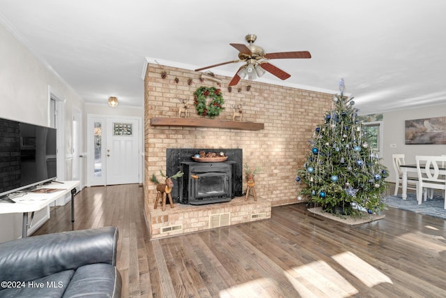 living room with a wood stove, hardwood / wood-style flooring, ceiling fan, ornamental molding, and brick wall