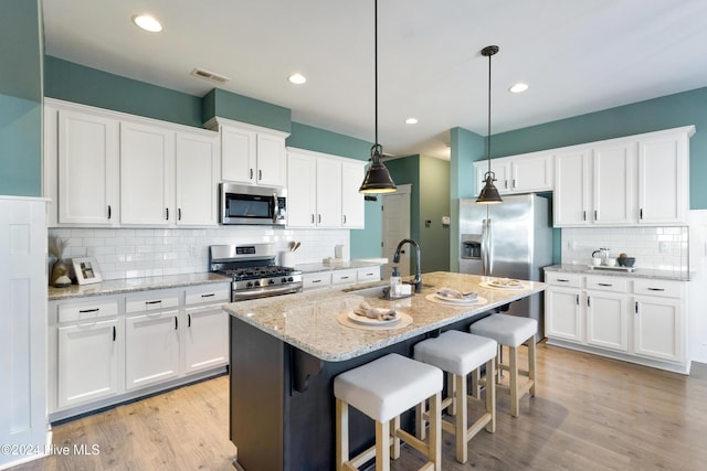 kitchen with appliances with stainless steel finishes, a kitchen island with sink, sink, white cabinets, and hanging light fixtures