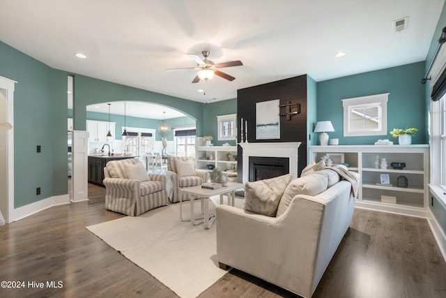 living room featuring ceiling fan with notable chandelier, a large fireplace, and dark wood-type flooring