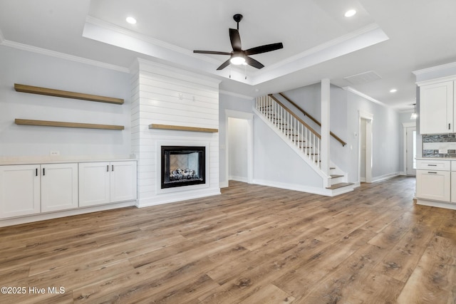 unfurnished living room featuring a tray ceiling, light wood-style floors, ornamental molding, a large fireplace, and stairs