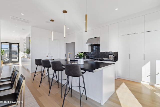 kitchen featuring light wood-type flooring, tasteful backsplash, decorative light fixtures, white cabinetry, and an island with sink