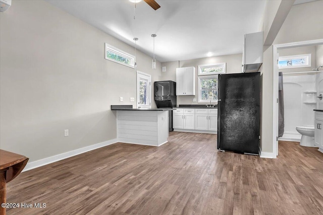 kitchen with wood-type flooring, hanging light fixtures, kitchen peninsula, black fridge, and white cabinetry