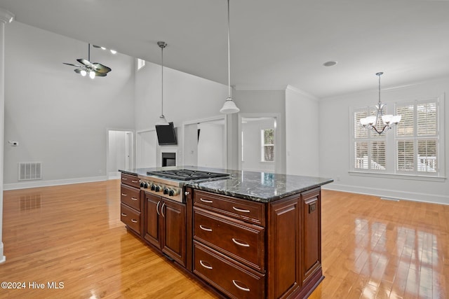 kitchen featuring stainless steel gas stovetop, ceiling fan with notable chandelier, light hardwood / wood-style flooring, dark stone countertops, and hanging light fixtures