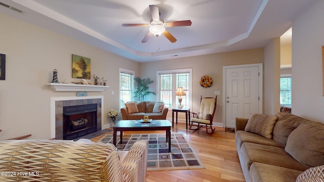 living room with a raised ceiling, a tiled fireplace, ceiling fan, and light hardwood / wood-style flooring