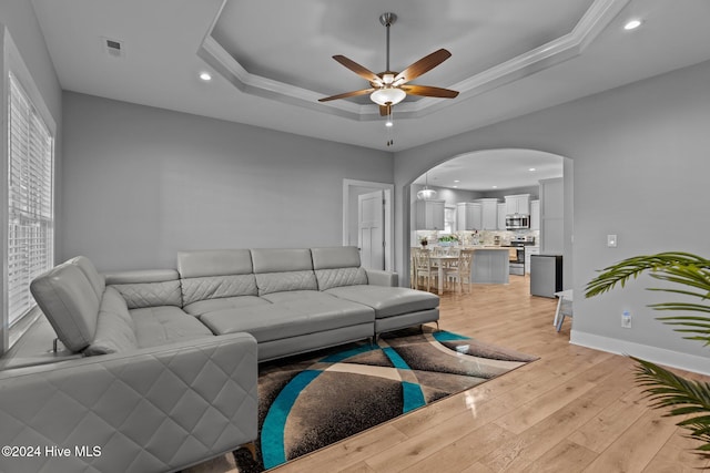 living room featuring ceiling fan, ornamental molding, a tray ceiling, and light hardwood / wood-style flooring