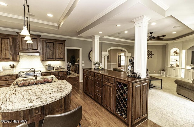 kitchen with a center island with sink, a tray ceiling, ornamental molding, dark brown cabinets, and decorative columns