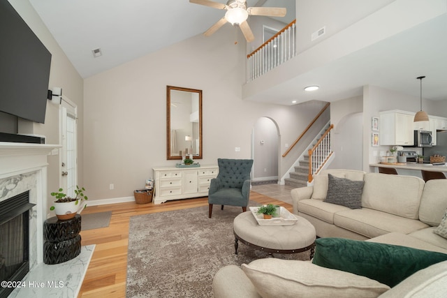 living room with high vaulted ceiling, ceiling fan, light wood-type flooring, and a high end fireplace